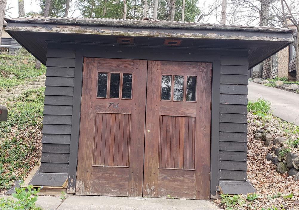 An image of a brown clapboard sided garage built into a hill. The garage doors are hinged like a standard door, and open in the middle.  This is image is on our About Humming Bird Woodworks page.