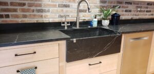 This image shows the Chicago Wright Soapstone sink, and soapstone countertops with the brick wall in the background. The birch wood base cabinets are to the Left of the dishwasher.