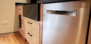 This image shows a closer view of the birch wood sink base cabinet with a drawer on top, The Garbage/Recycling drawer beneath. To the right of that is two large drawers beneath the sink.  Rge dishwasher is on the right.