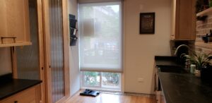 This image shows part of the Fridge wall on the left. To the right of those cabinets there is a doorway that leads to the front hall. That wood pocket door has a fluted glass panel. To the right of the pocket door is a wood slated wall with a fluted glass panel attached to it that can be opened. On the far right of the image is the sink side of the kitchen.