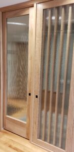 This image shows in closer detail that wood pocket door has a fluted glass panel. To the right of the pocket door is a wood slated wall with a fluted glass panel attached to it. This panel can be opened. Here is is shown in the closed position.