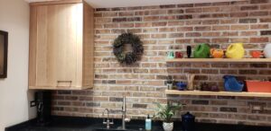 This image shows the single door upper Birch wood cabinet on the Left.  The 2 Birch wood floating shelves on the right and a decorative wreath hanging above the sink on the brick wa