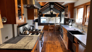 This image shows the full kitchen looking East with the range side on the Left, and Sink side on the Right. At the far end of the kitchen are three plate glass window that overlook an outdoor shelter. The ceiling in this area is vaulted. The vaulted ceiling caries out into the outdoor shelter. Glen Ellyn Hilltop Gallery