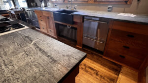 This image shows the Birch wood base cabinets on the sink wall side. The under counter drawer Fridge and Freezers on the far Left near outdoor shelter. To the right is a base cabinet with a bank of drawers on the left, and to the right of the bank of drawers. The smaller drawer on the Left matches the width of the Recycling and Garbage pull-out drawer with a shallow drawer beneath it. These two are set back 2" back from the upper drawer at the top. Around the sink there is a removable front to access the radiator beneath the sink. to the right of the sink there is a Double Dishdrawer. Next to the Dishdrawers and far right are a bank of 4 drawers with the top drawer 2" out from the lower three drawers. Glen Ellyn Hilltop Gallery