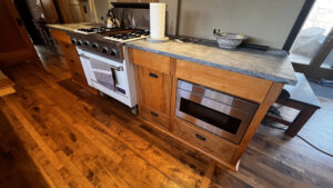 This image shows the lower cabinets on the range side of the kitchen. In the cabinet to the left of the range there is one top drawer that extends the whole width of the cabinet. Beneath that drawer set 2" back on the Left in this cabinet are 3 stacked drawers. In this cabinet on the right is a tray storage compartment that is the height of the tree lower drawers. The cabinet to the right holds 2 upper drawers on top. The smaller drawer on the Left matches the width of the Recycling and Garbage pull-out drawer with a shallow drawer beneath it. These two are set back 2" back from the upper drawer at the top. There are two wider drawers to the right of the Recycling drawer. There is one drawer at the top with a drawer microwave set 2" back beneath the top drawer. Beneath that is a wider shallow drawer. There are Soapstone countertops on top of these cabinets. Glen Ellyn Hilltop Gallery