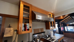 This image shows another angle of the single door Birch wood cabinet with Glass panel insert on the Left. 48" range hood in the center with decorative wood panel above. To the right is a double door Birch cabinet with with Glass panel insert on the right. Glen Ellyn Hilltop Gallery