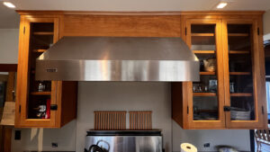 This image shows The single door Birch wood cabinet with Glass panel insert on the Left. 48" range hood in the center with decorative wood panel above. To the right is a double door Birch cabinet with with Glass panel insert on the right. Glen Ellyn Hilltop Gallery