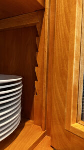 This image shows a Birch wood cabinet with wood saw-tooth shelf adjusters, and wood adjustable shelf supports. There twelve white dinner plates stacked on the bottom shelf on the left side of the image. The stile and rail cabinet door with glass insert is partially opened.