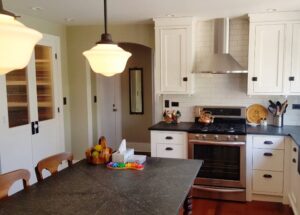 This image shows the overall view of the stove wall elevation. The kitchen cabinets are made from poplar wood and painted white. The chimney hood is on a white subway tile wall with the stove below.