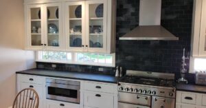 This image shows a poplar wood double upper cabinet with glass paneled doors. Directly below are transom windows above the soapstone countertops. Below the counters are a set of drawers surrounding a drawer microwave. The AGA Stove and Chimney hood are on the right