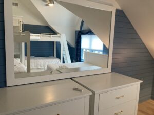 This image shows the two bunk beds with four beds Bunk Beds painted white reflected in the mirror over two dressers. It is in and L shaped configuration with the beds coming out of a central tower. The walls are painted a dark blue on the lower walls with white above.