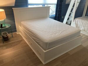 The image shows the queen sized bed with head board painted white. There are four drawers on the side of the bed frame beneath the Queen sized mattress. Humming Bird Exclusives.