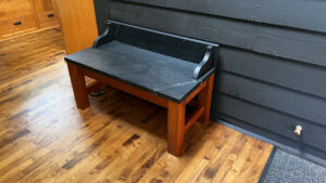 This image shows the wood base warming bench with soapstone top against an interior clapboard sided wall. This view is showing the Right side, and front view of the bench.