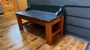 This image shows the a closer up view of the wood base warming bench with soapstone top against an interior clapboard sided wall. This view is showing the Right side, and front view of the bench. 
Humming Bird Exclusives.