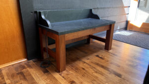 This image shows the a closer up view view of the wood base warming bench with soapstone top against an interior clapboard sided wall. This view is showing the Left side, and front view of the bench.