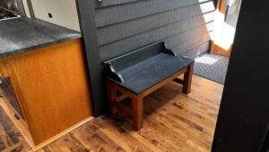 This image shows the wood base warming bench with soapstone top against an interior clapboard sided wall. This view is showing the Left side, and front view of the bench. Humming Bird Exclusives.